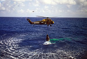 A green helicopter hovers low over the water, with the Mercury capsule suspended below. The helicopter has "Marines" written on it, and the number "44"