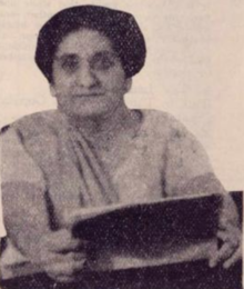 An older South Asian woman wearing light-colored sari and holding an open book in two hands