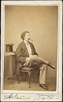 Photographie d'Artemus Ward, assis avec sa jambe droite croisée sur sa gauche, un haut-de-forme et des livres posés sur la table à sa droite.  Une légende dactylographiée au bas de l'image lit "H. Hering Photo" et son nom est écrit en bas.