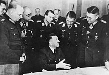 Photographie en noir et blanc de Hitler, assis au centre de l'image, entouré d'officiers supérieurs au quartier général du groupe d'armées Vistule en mars 1945