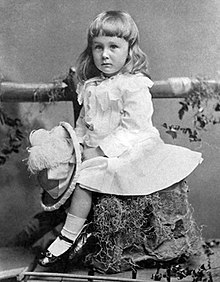 A young boy sits on a tree stump. He holds a large feathered hat in his hand.