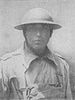 Head and shoulders of a man in a military coat, buttoned up to the neck, with a light pack strapped to his chest. He is wearing a metal helmet with a wide brim, its chin strap tightly cinched.