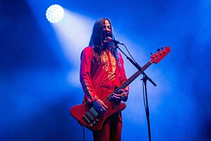 Man with shoulder length hair holding playing a bass guitar and singing into a microphone