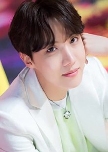 J-Hope in a white suit against a yellow, purple, and red background, his left hand rests on his temple
