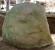 An eroded sculpture carved in relief onto a boulder. It shows a human figure from the front, squatting with the legs splayed. Its elbows are doubled and under each arm it grips a barely distinguishable animal. The figure's face is mostly eroded away but hollows for the eyes and mouth are still visible. The figure wears large ear-spools and a prominent headdress.
