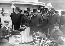 Groupe d'hommes portant des chapeaux melons et des redingotes sur le pont d'un navire devant les membres d'un orchestre