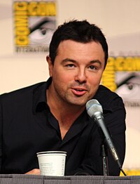 A man with black hair and a black shirt, leans forward slightly to speak into a microphone.