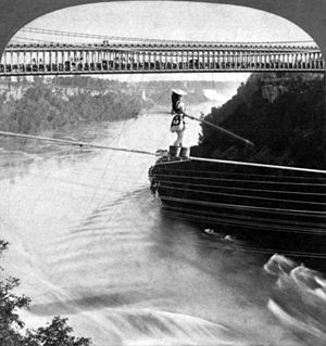 A woman is crossing on a rope over a river. She wears a wide-brim hat and holds a pole to balance herself while her feet are in buckets. A double-deck bridge, filled with audience on the lower deck, is in the background.