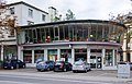 Trier, Theodor-Heuss-Allee 18: der weitgehend in große Fenster aufgelöste Eckladen, 1933 durch Architekturbüro Brand und Mertes einem Wohnhaus von 1880 angebaut, 1956 zeittypisch aufgestockt mit polygonaler Verglasung mit schräg nach außen gestellter Fenstern