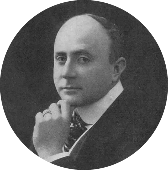 head and shoulders of balding white man in early middle age, clean-shaven, in lounge suit and tie, semi-profile, looking towards the camera