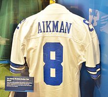 Le dos d'un maillot blanc derrière une vitrine, avec l'inscription Aikman en lettres bleues au-dessus du numéro 8 bleu.