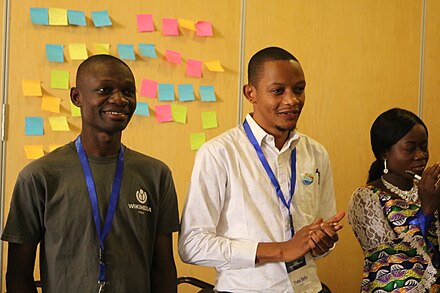 3 Participants in front of a wall of red, blue, green and blue sticky notes. They are wearing lanyards with their names and one of them is clapping.