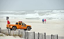 Пикап Pensacola Beach Lifeguard припаркован на пляже с бурным морем.  Два человека идут близко к кромке воды на заднем плане и еще двое идут к воде в центре изображения.