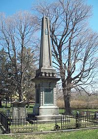Каменный обелиск и надгробие внутри невысокой черной ограды посреди кладбища. Обелиск стоит на декоративном квадратном постаменте с металлической табличкой зеленого цвета на нем.