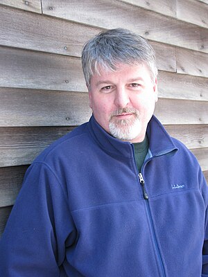 A portrait photograph of a white man in a blue jacket; he has greying hair on both his head and face and is looking into the camera with a neutral expression.