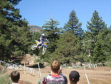 Moto en pleine course photographiée dans les airs, sur piste campagnarde ; la photo comprend trois personnes caucasiennes de dos au premier plan.