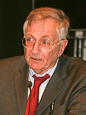 Head and shoulder photo of older white man wearing wire-rimmed glasses, a brown suit and red tie; he is speaking into a microphone