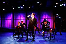 Image of brown-haired man dressed in a navy and red school uniform singing into a microphone while surrounded by other ensemble members, also dressed in school uniforms