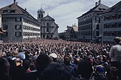 Fotografia de uma multidão em uma praça pública, olhando para uma plataforma;  no fundo uma igreja.