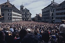 Photographie d'une foule sur une place publique, regardant une estrade ; au fond une église.