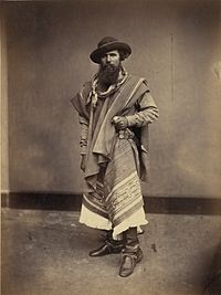 Full-length photograph of a bearded man wearing a wide-brimmed hat, bandana, serape, leather boots and grasping the handle of a long knife in his left hand