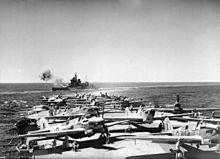 Photo en noir et blanc du pont d'un porte-avions couvert d'avions, un autre bateau à l'arrière-plan.