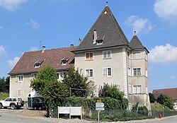 A grey building, in two sections, one two storeys tall with tall, angled, brown tiled roof, the other two intersecting square towers, three storeys tall, each with tall square-pyramidal slate roofs.