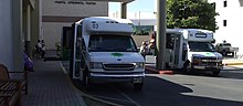 Two Maui Bus buses engaged in paratransit services. One is picking up a person who uses a wheelchair, while another is discharging another person using a mobility scooter. MauiBusParatransitCrop.JPG