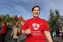 A woman wearing a Labour Party t-shirt with Sámi text on it. On the top right side of the shit are two pins, one pin has the logo of the Workers' Youth League logo. The pin next to it has the word 'Frihet' which translates to 'Freedom'