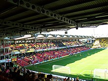 Tribun dua tingkat dengan bangku berwarna merah, kuning, dan hitam, berada di samping lapangan sepak bola.