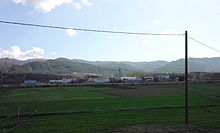Au fond de la photo, une petite zone artisanale, avec quelques hangars métalliques ; au premier plan, grand champ verdoyant et poteaux téléphoniques. En arrière-plan, des collines.