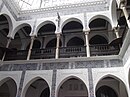 A photograph of a structure with two floors, only the top of the lower floor and some of the roof is pictured. There are ornate designs on the walls, arches, columns and railings