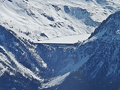 Photographie d'un barrage enneigé en hiver en contre-plongée, tout est de neige : le barrage est blanc, l'eau glacée étant recouverte par une fine couche de neige.