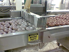 Maschinelles Überziehen von Donuts mit einer Zuckerglasur (hier bei Massenherstellung in einer Krispy-Kreme-Filiale in den USA)