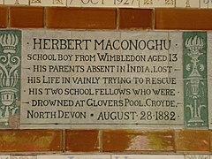 Een tablet gevormd uit zes tegels van standaardformaat, omzoomd door groene bloemen in de stijl van de Arts and Crafts-beweging.  Op het tablet staat: "Herbert Maconoghu schooljongen uit Wimbledon, 13 jaar oud. Zijn ouders, die afwezig waren in India, verloren zijn leven door tevergeefs zijn twee schoolgenoten te redden die verdronken waren in Glovers Pool, Croyde, North Devon, 28 augustus 1882".