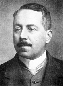 Portrait of a moustached Tanner in a suit with waistcoat and necktie, facing slightly to the left. He has combed over hair and his right eyebrow is slightly raised.