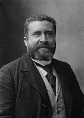 Retrato en blanco y negro de un hombre con canas, bigote y perilla, vestido con corbata, camisa blanca, chaqueta clara y chaqueta oscura