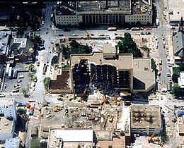Image aérienne d'un bâtiment détruit, noircit, ouvert sur l'un de ses côtés, où des secours travaillent sur un champ de gravats.