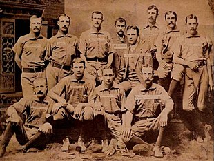 A sepia photograph of twelve men arranged in two rows, standing and sitting. Ten are wearing light baseball uniforms with with dark socks, while two are dressed in suits.