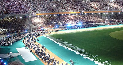 In the foreground is a field with a large tree in the middle. The tree appears to be real, but is situated on what is clearly a temporary structure. Dozens of people are standing around the edge of the field, while thousands are watching from a full, brightly illuminated covered seating area beyond the perimeter.