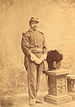 Retrato de corpo inteiro de um homem negro com bigode em um uniforme militar ornamentado, incluindo luvas brancas, ombreiras, boné e uma espada fina amarrada na cintura.  Ao lado dele está um pedestal com um capacete militar cerimonial no topo.