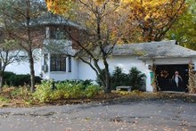 A small white brick gatehouse in front a forest of changing leaves, with a black asphalt driveway on its left