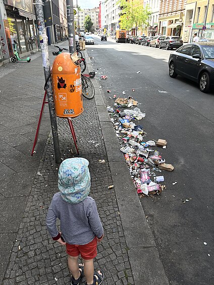 The morning after the carnival of cultures party in Berlin Kreuzberg (Germany): A local child looking at the trash on the streets and an orange machine from the BSR cleaning up.