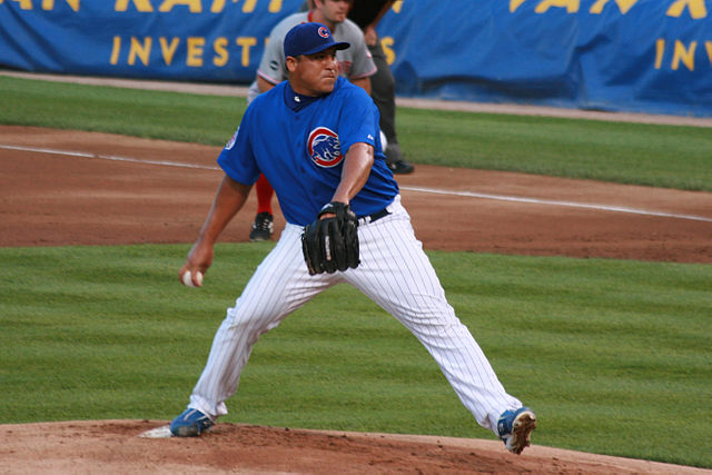 Full shot of pitcher in mid pitch.  Pitcher is wearing a blue hat, a blue shirt with a Cubs logo and white pants