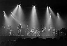 Image monochrome de Pink Floyd se produisant sur une scène de concert. Chaque membre du groupe est éclairé par le haut par des projecteurs lumineux.