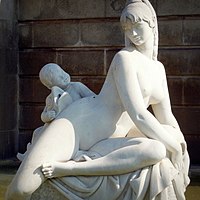 Escultura de piedra blanca que representa a una mujer sentada, recostada ligeramente sobre su lado izquierdo y un niño tras ella acodado en el muslo de la mujer y la cabeza descansando sobre el puño. Ambos desnudos, se miran la cara