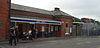 A brown-bricked building with a blue sign reading "EPPING STATION" in white letters and people in the foreground all under a blue sky with white clouds