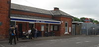 A brown-bricked building with a blue sign reading "EPPING STATION" in white letters and people in the foreground all under a blue sky with white clouds