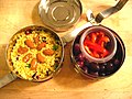 Différents éléments qui composent une gamelle ronde à plateaux ; dans un des plateaux, du riz safrané est décoré d’amandes sèches, dans l’autre un ravier en verre (ou en plastique) qui contient des lamelles de poivron rouge est déposé sur un lit d’olives noires.