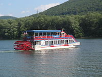 A red, white and blue paddlewheel boat with a sign "Hiawatha" on a river with a forested mountain behind